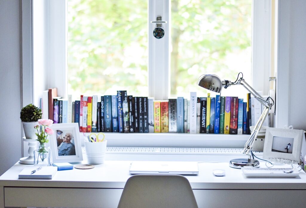 A white desk by a tall window with books along the sill and tree branches outside