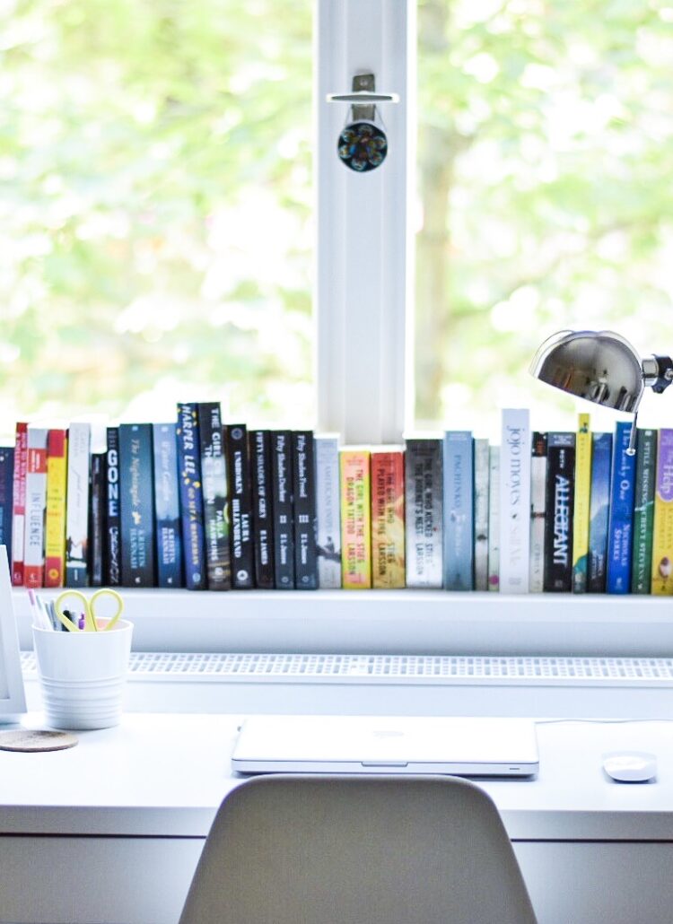 A white desk by a tall window with books along the sill and tree branches outside