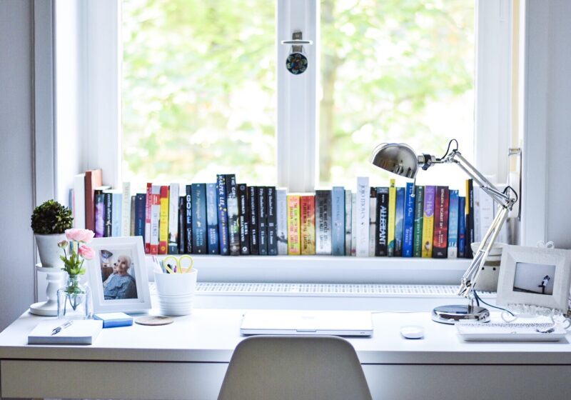 A white desk by a tall window with books along the sill and tree branches outside